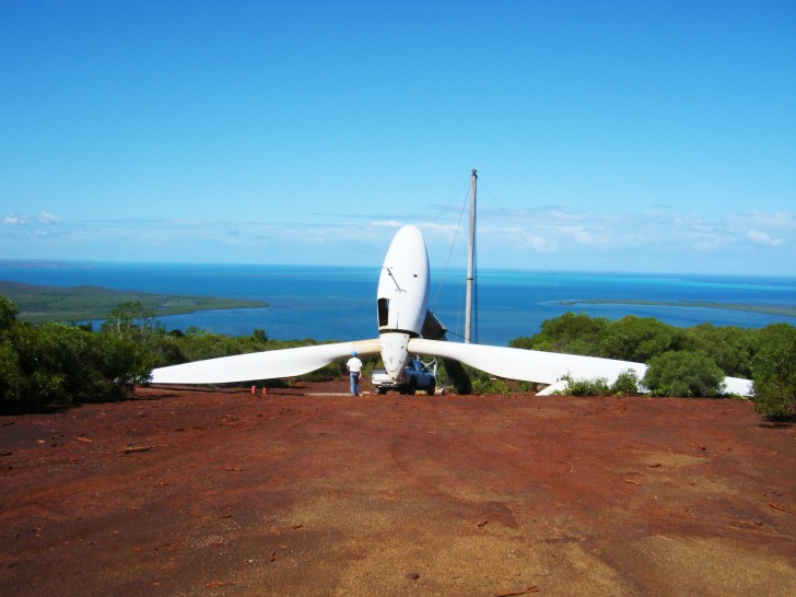 Bauarbeiter errichten eine Windkraftanlage auf Neukaledonien. Im Vordergrund sieht man rote Erde und eine liegende Windkraftanlage, im Hintergrund grüne Vegetation und das türkisblaue Meer, sowie blauen Himmel.